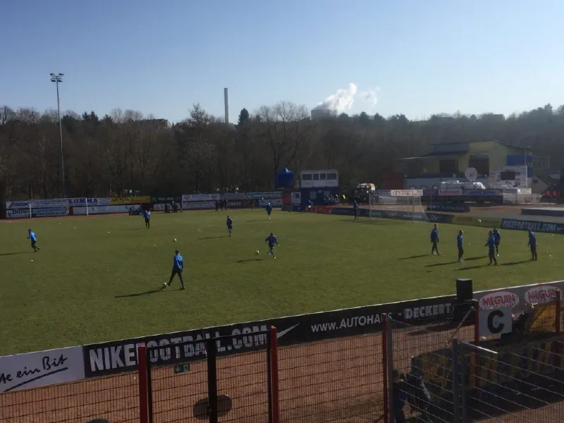 Spieler des 1. FC Saarbrücken in blauen Trikots beim Aufwärmen auf dem Rasen, dahinter Werbebanden und ein dampfender Industrieschornstein.